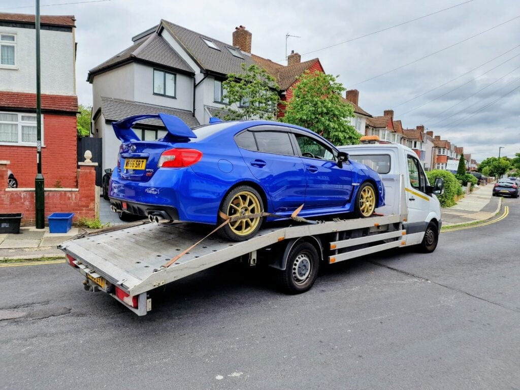 Flatbed tow truck transporting a blue Subaru on a London residential street — car recovery London.