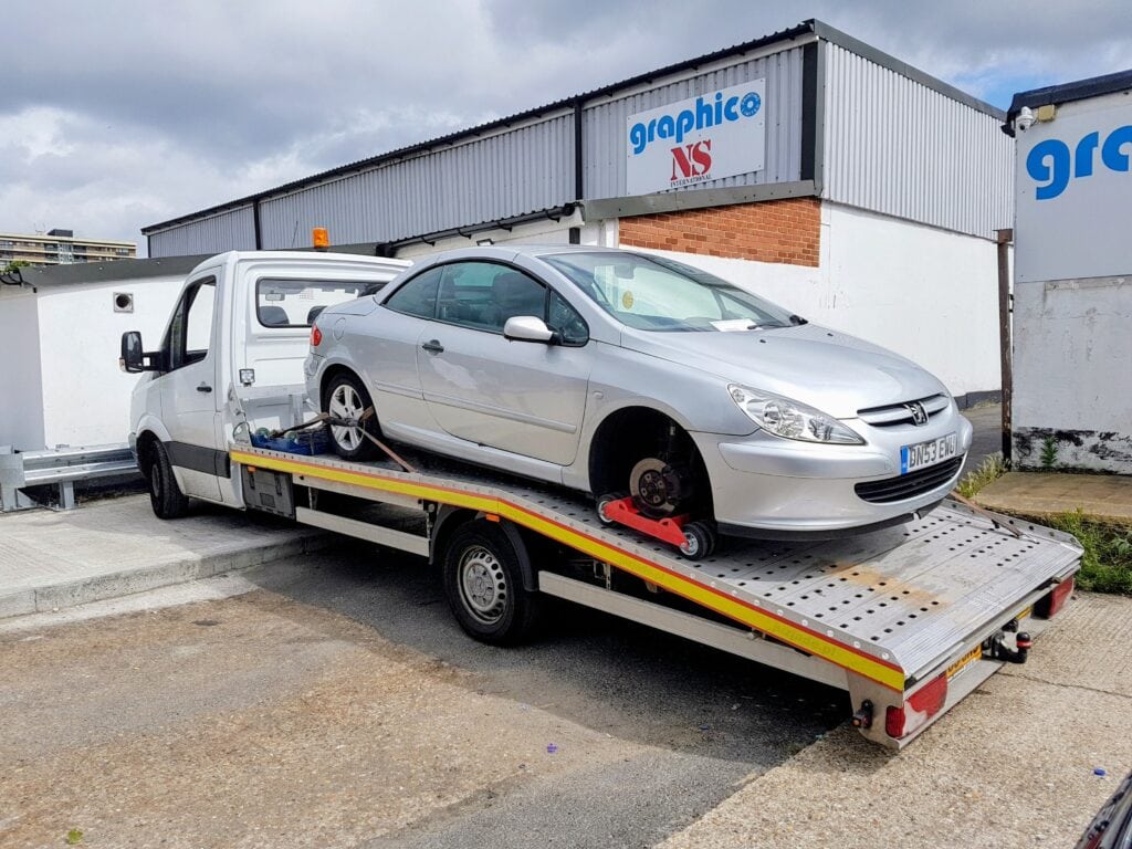 Scrap car collection London — flatbed tow truck loading a silver Peugeot with a missing wheel