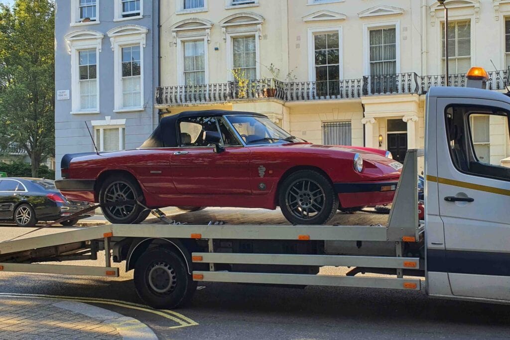 A classic red Alfa Romeo Spider securely loaded onto a flatbed recovery truck on a residential street, showcasing reliable breakdown recovery.