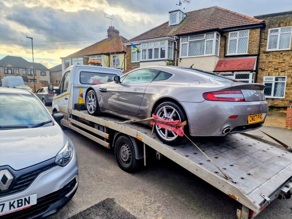 Silver Aston Martin V8 Vantage loaded onto a recovery truck for emergency breakdown service in a London residential area.