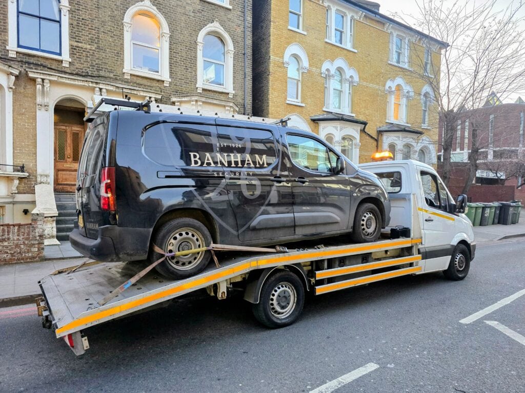 A black Banham Security van secured on a flatbed recovery truck on a residential street with terraced houses.