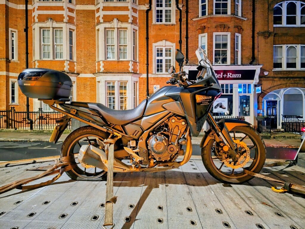 A Banham Security Honda patrol motorcycle secured on a flatbed recovery truck at night on a London street with red brick buildings.