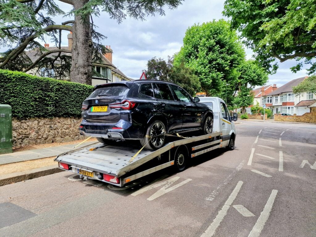 A black BMW X3 SUV secured on a flatbed recovery truck on a tree-lined residential street in London.