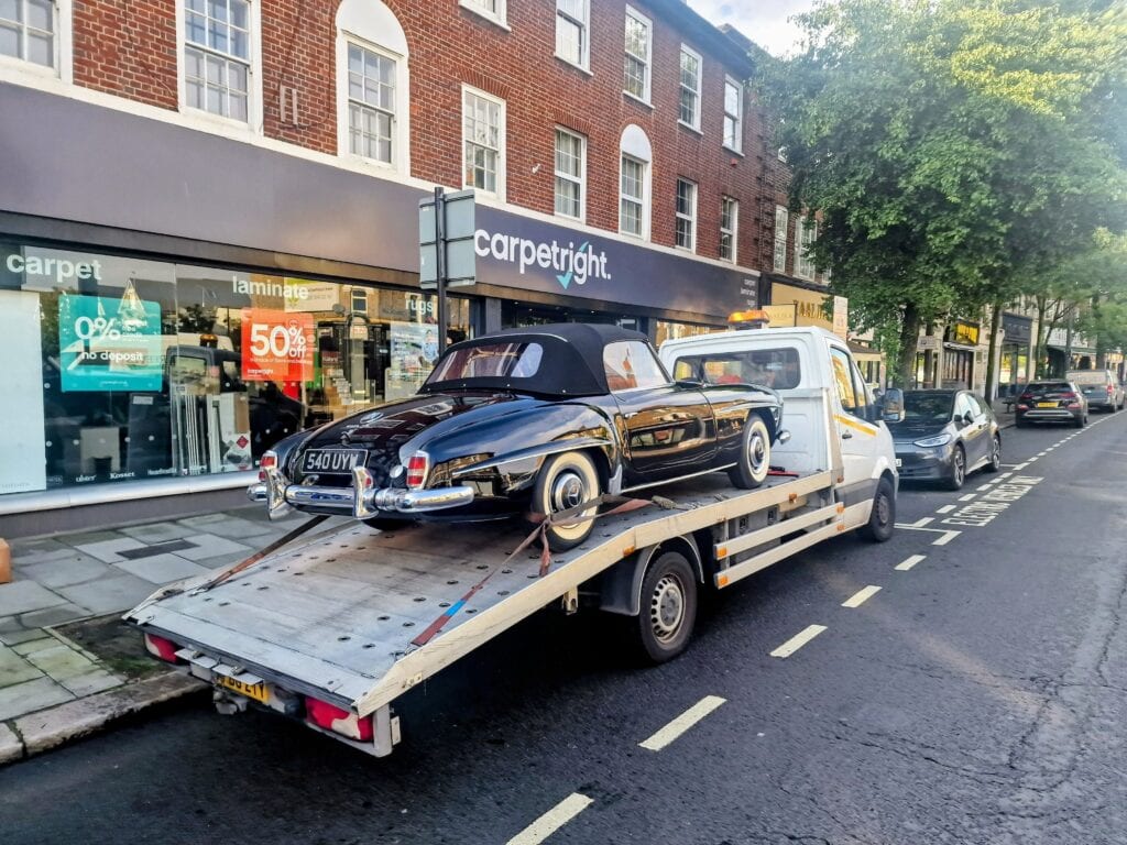A rare black Mercedes-Benz 190 SL convertible with whitewall tires secured on a flatbed recovery truck on a high street with shops.