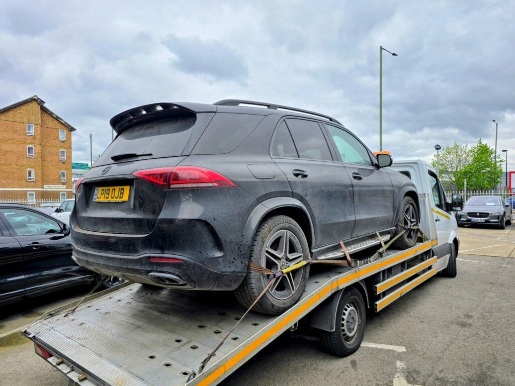 A black Mercedes-Benz GLE SUV being transported on a flatbed recovery truck in a London car park.