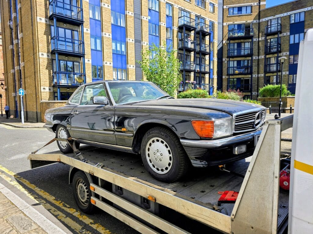 A classic black Mercedes-Benz R107 SL roadster being transported on a flatbed recovery truck through a residential area in London.