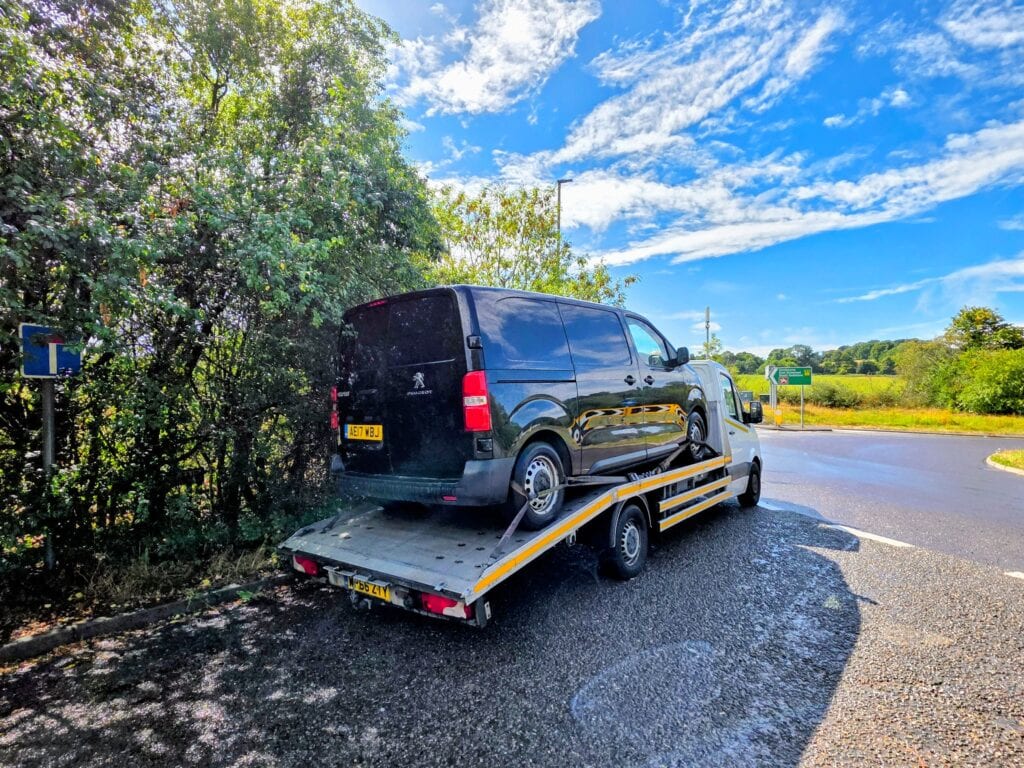 A black Peugeot Traveller MPV secured on a flatbed recovery truck near a country roundabout with green trees.