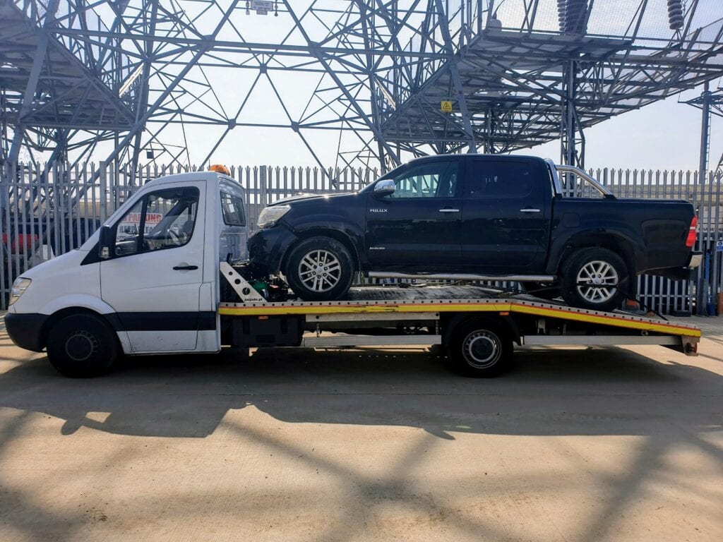 A black Toyota Hilux double cab pickup truck secured on a flatbed recovery truck in an industrial area with large electrical pylons in the background.