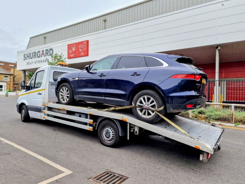 A dark blue Jaguar F-Pace SUV secured on a flatbed recovery truck outside a Shurgard Self-Storage facility.