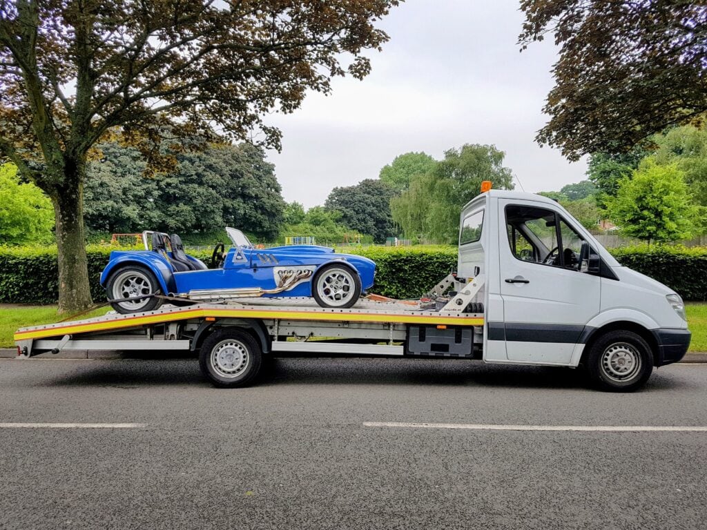 A blue open-top kit car with a chrome side exhaust secured on a flatbed recovery truck against a background of green trees.