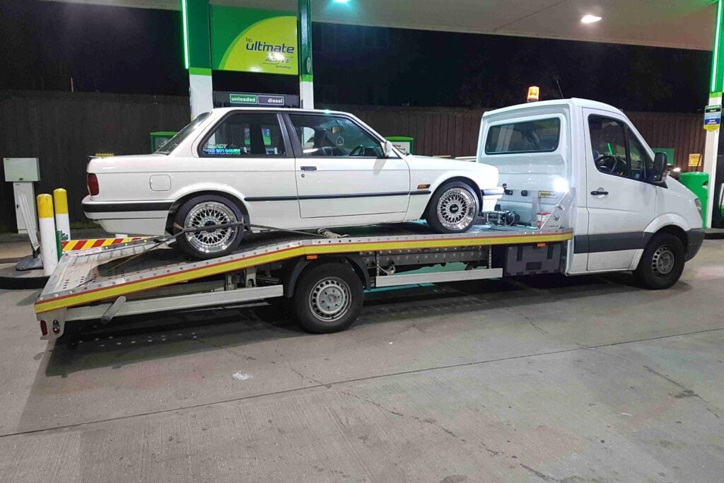 A white modified BMW E30 coupe with custom wheels securely loaded onto a flatbed recovery truck at a petrol station at night.