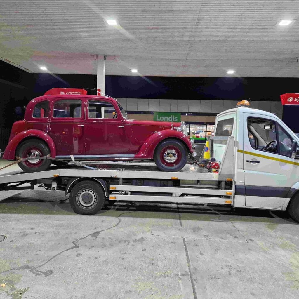 Classic car loaded on flatbed tow truck at London petrol station, ready for careful recovery.