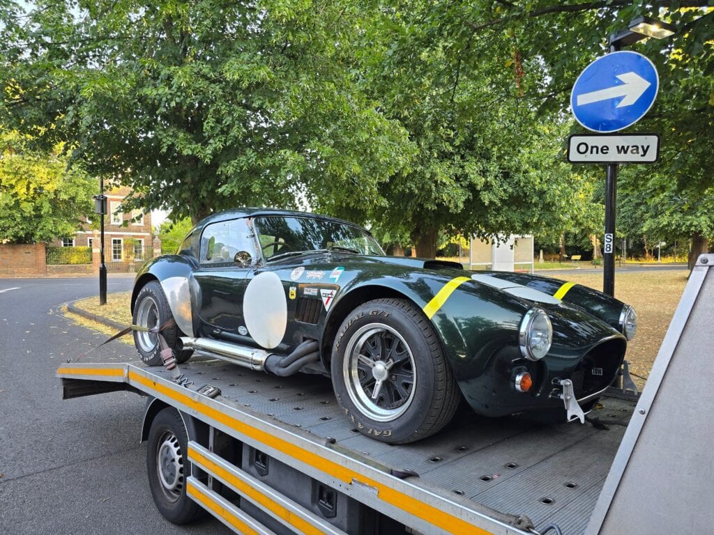 A dark green AC Cobra with a racing hardtop and yellow stripes securely strapped to a flatbed recovery truck, highlighting specialist vehicle transport.