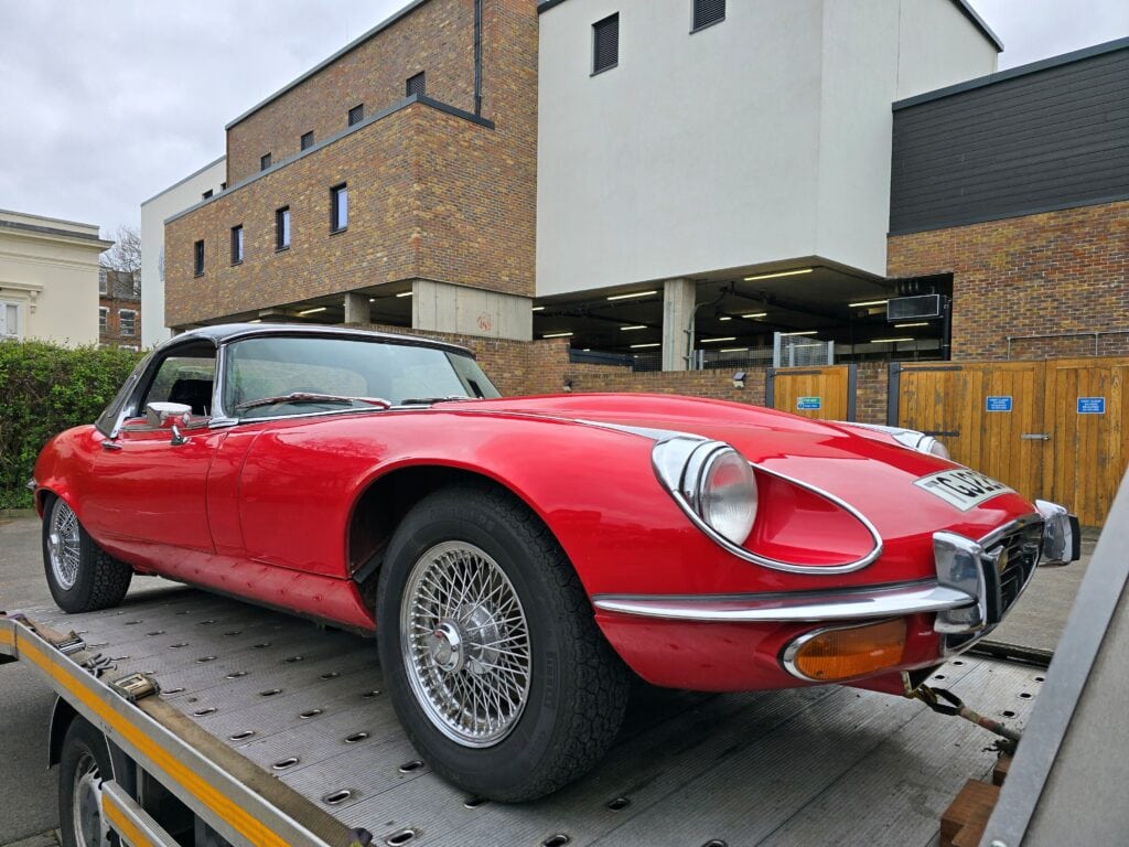 A classic red Jaguar E-Type Series 3 V12 securely strapped to a flatbed recovery truck near a modern building, demonstrating careful vehicle transport.