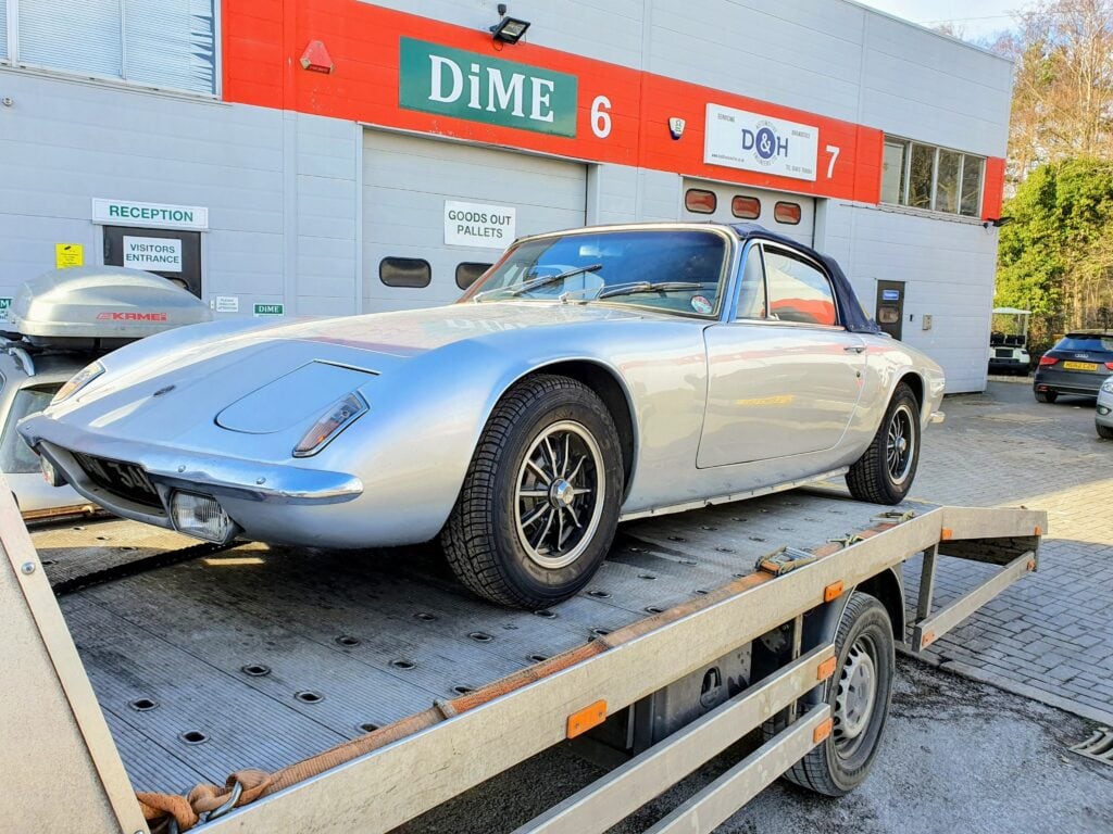A silver vintage Lotus Elan +2 securely loaded onto a flatbed recovery truck outside a specialist garage, showcasing expert classic car recovery.