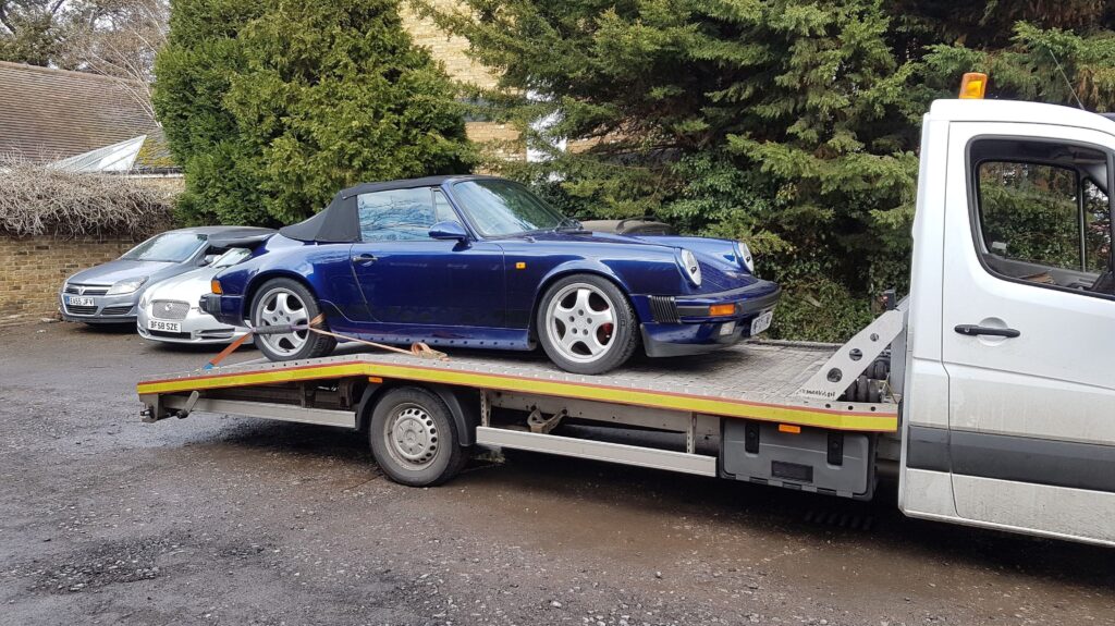 A dark blue vintage Porsche 911 Cabriolet securely loaded onto a flatbed recovery truck, demonstrating safe transport for classic sports cars.