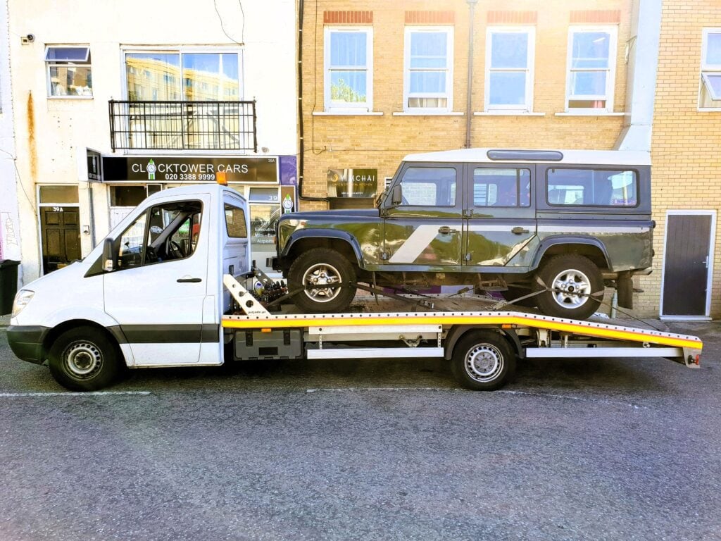 A classic dark green Land Rover Defender 110 secured on a flatbed recovery truck on a high street outside local shops.