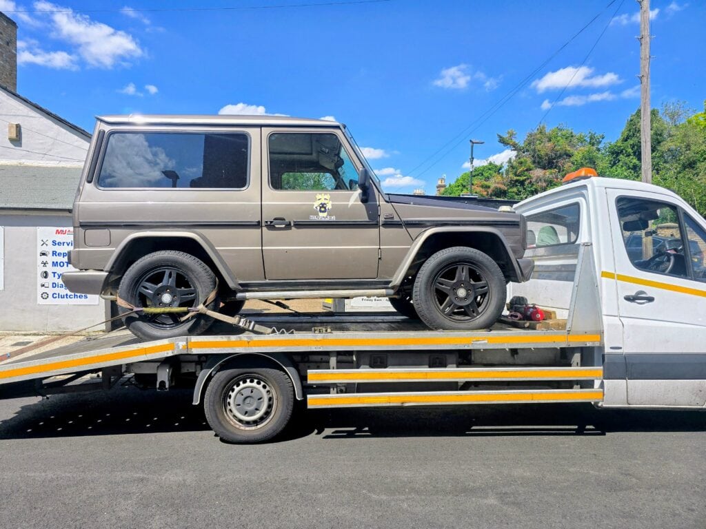 A classic bronze Mercedes-Benz G-Wagon short wheelbase securely loaded onto a flatbed recovery truck on a sunny day in Surrey.