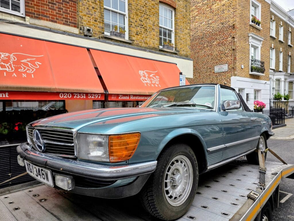 A classic metallic blue Mercedes-Benz R107 SL convertible being transported on a flatbed recovery truck outside a restaurant in London.