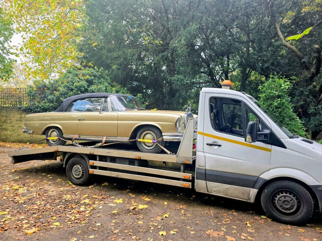 A gold classic Mercedes-Benz W111 Coupe barn find being transported on a flatbed recovery truck in a wooded area of Surrey.