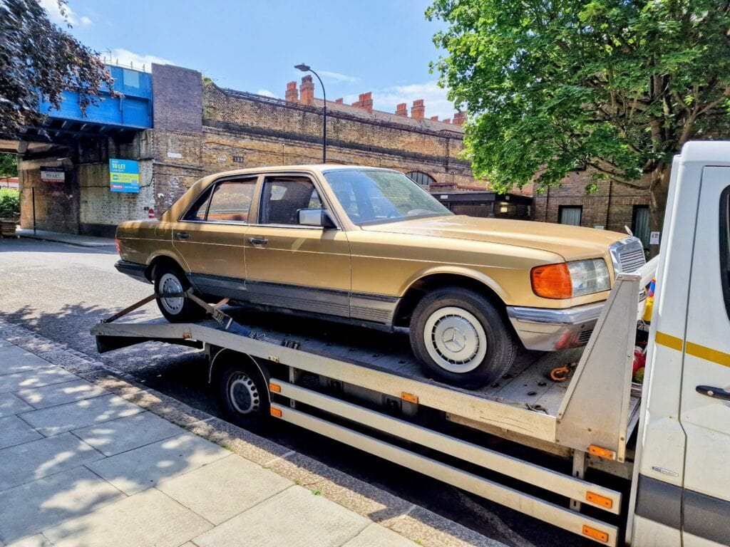 A classic gold Mercedes-Benz W126 S-Class sedan securely loaded onto a flatbed recovery truck on a city street in London.