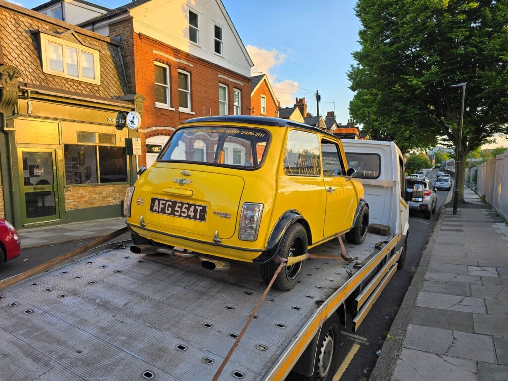 A bright yellow classic Mini Cooper with an "Italian Job" sticker securely loaded onto a flatbed truck on a London residential street.