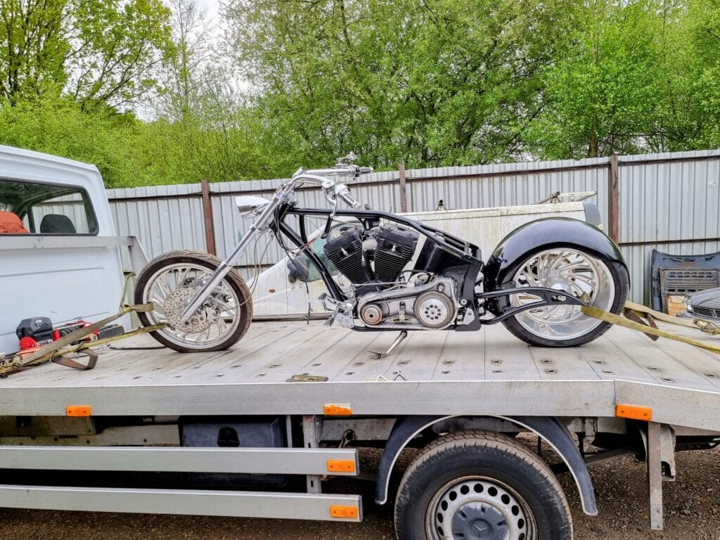 A custom chopper motorcycle rolling chassis with a V-twin engine and wide rear wheel secured on a flatbed recovery truck.