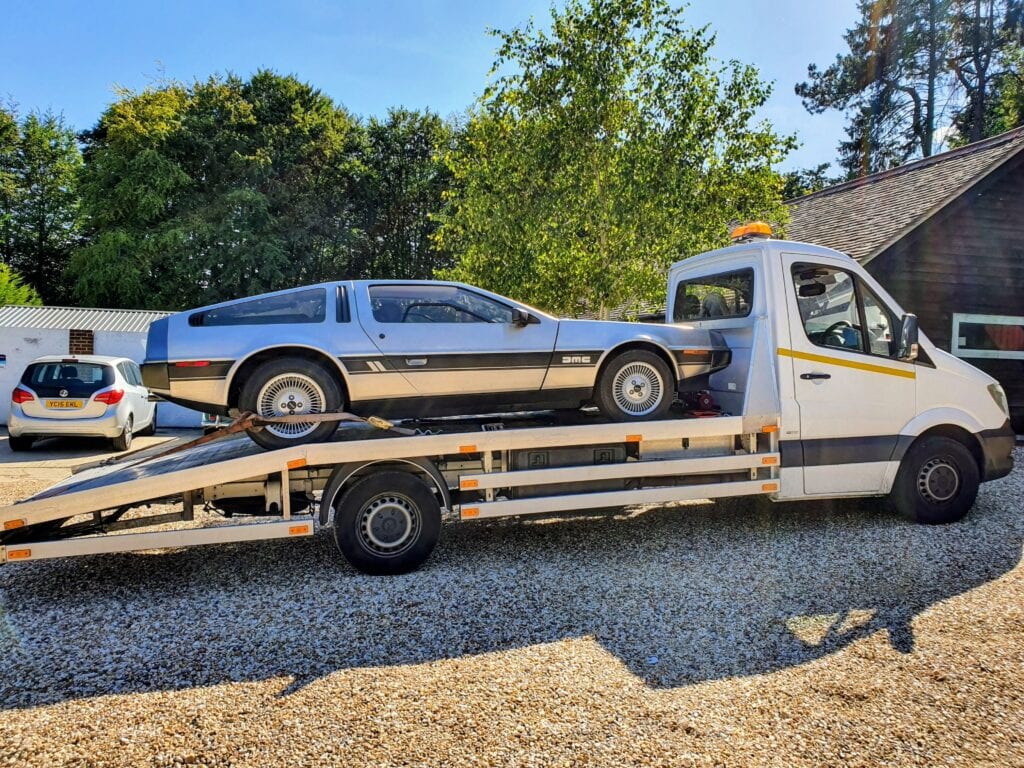 DeLorean DMC-12 with gull-wing doors open secured on a flatbed recovery truck in a sunny driveway.