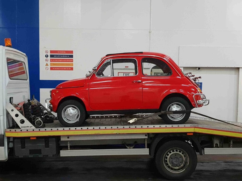 A classic red Fiat 500 securely loaded onto a flatbed recovery truck on a cross-channel ferry, demonstrating European vehicle collection.