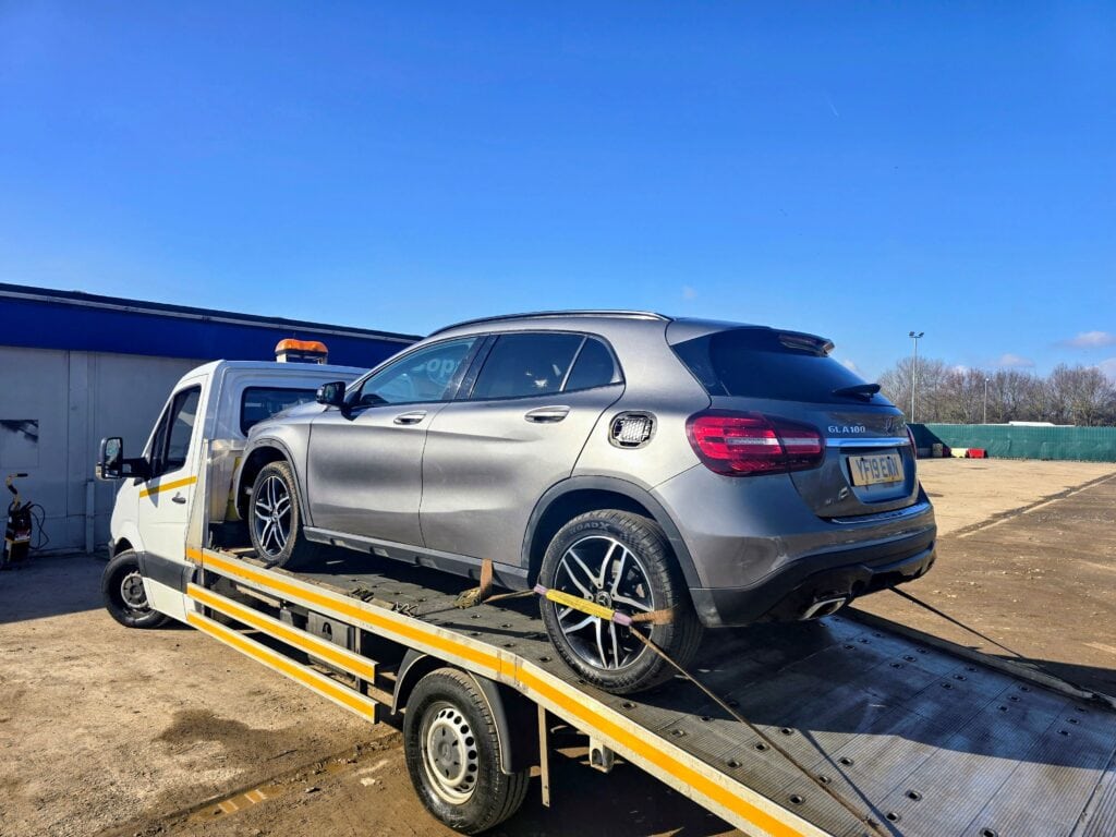 A grey Mercedes-Benz GLA being collected from a Copart auction site and loaded onto a flatbed recovery truck under a clear blue sky.