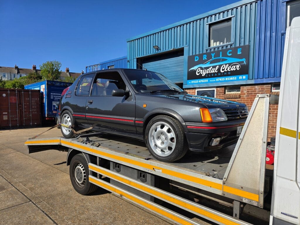 A classic grey Peugeot 205 GT secured on a flatbed recovery truck outside a detailing specialist unit in London.