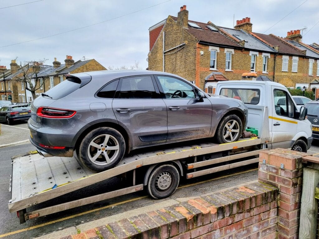 A grey Porsche Macan SUV secured on a flatbed recovery truck on a residential street with brick terraced houses.