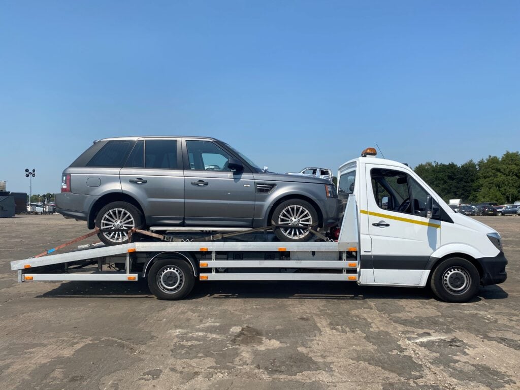 A grey Range Rover Sport SUV secured on a flatbed recovery truck in a large open vehicle compound.