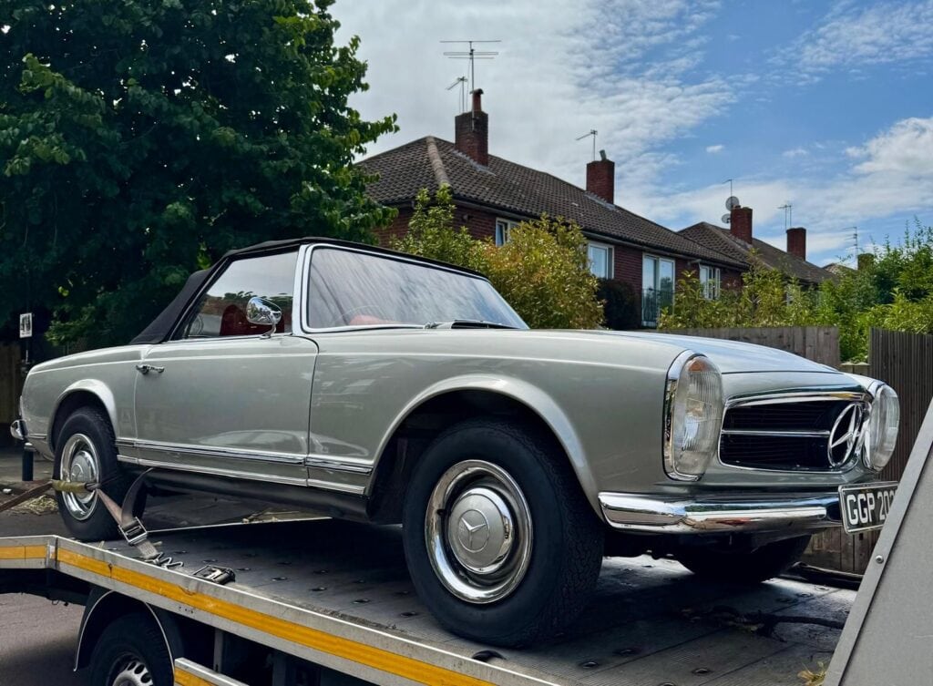 A vintage silver Mercedes-Benz Pagoda W113 securely loaded onto a flatbed recovery truck on a residential street in London.