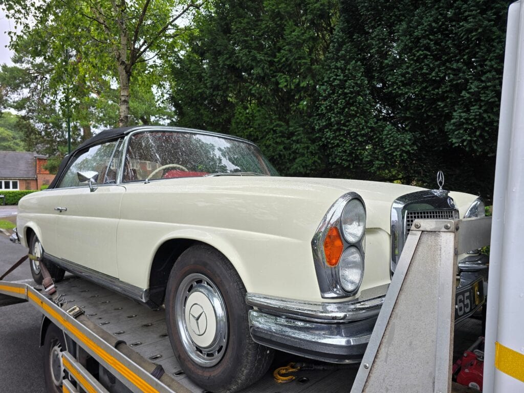 A classic ivory Mercedes-Benz W111 Cabriolet secured on a flatbed recovery truck in a green London street.
