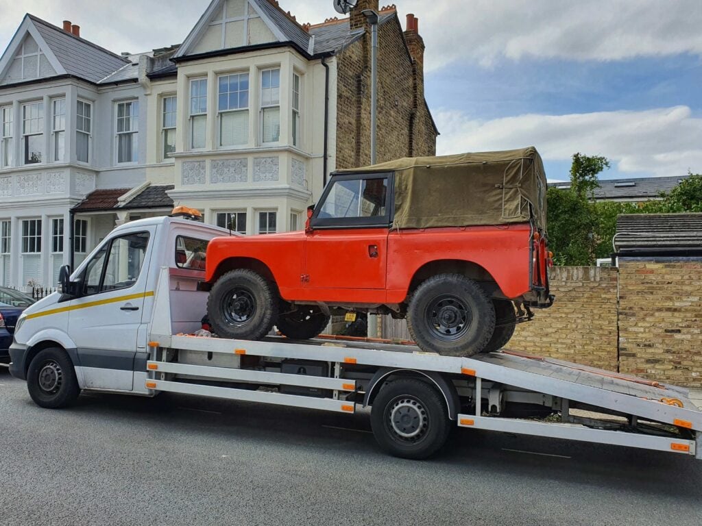 A vintage orange Land Rover Series 2A with a canvas roof securely loaded onto a flatbed recovery truck on a residential street.