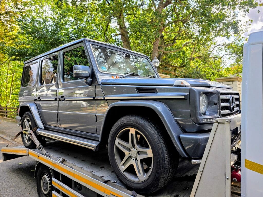 A grey Mercedes-Benz G-Class AMG SUV securely loaded onto a flatbed tow truck on a tree-lined road in Surrey, following a mechanical breakdown.