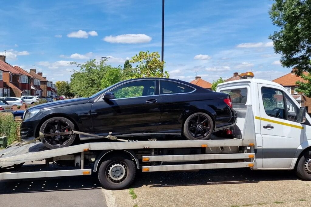 A black Mercedes-Benz C-Class Coupe securely loaded onto a flatbed recovery truck in a residential street in London.