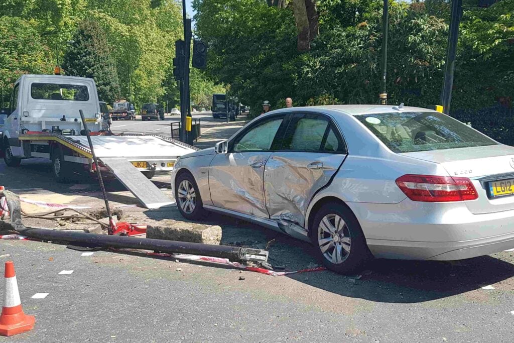 A silver Mercedes-Benz E-Class with heavy side impact damage near a fallen traffic light on a London street.
