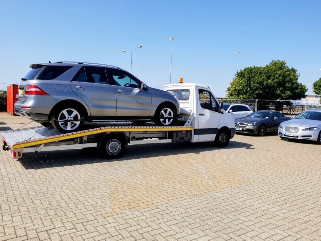 A silver Mercedes-Benz ML SUV securely loaded onto a flatbed recovery truck at a dealership compound.