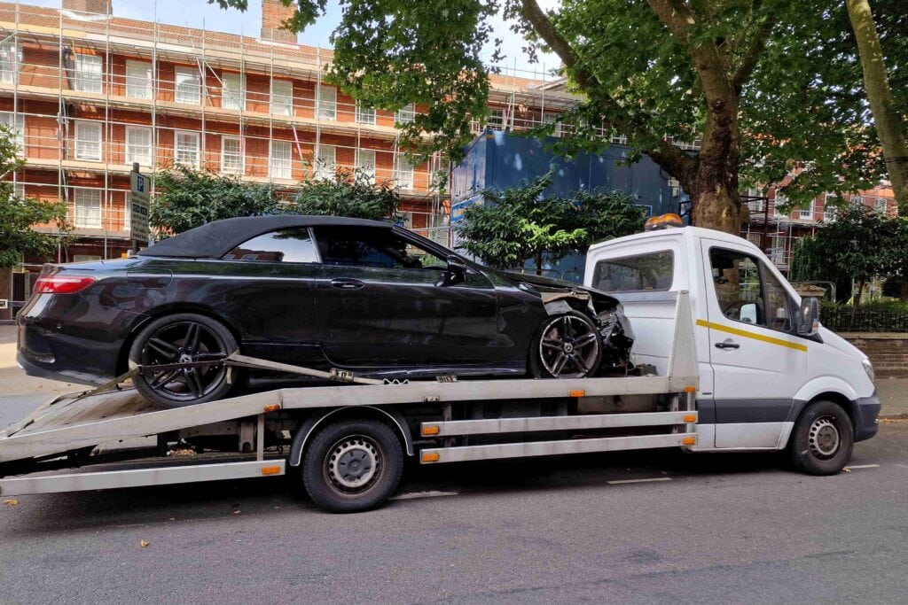 A black Mercedes-Benz S-Class Convertible with heavy front-right damage securely loaded onto a flatbed recovery truck on a London street.
