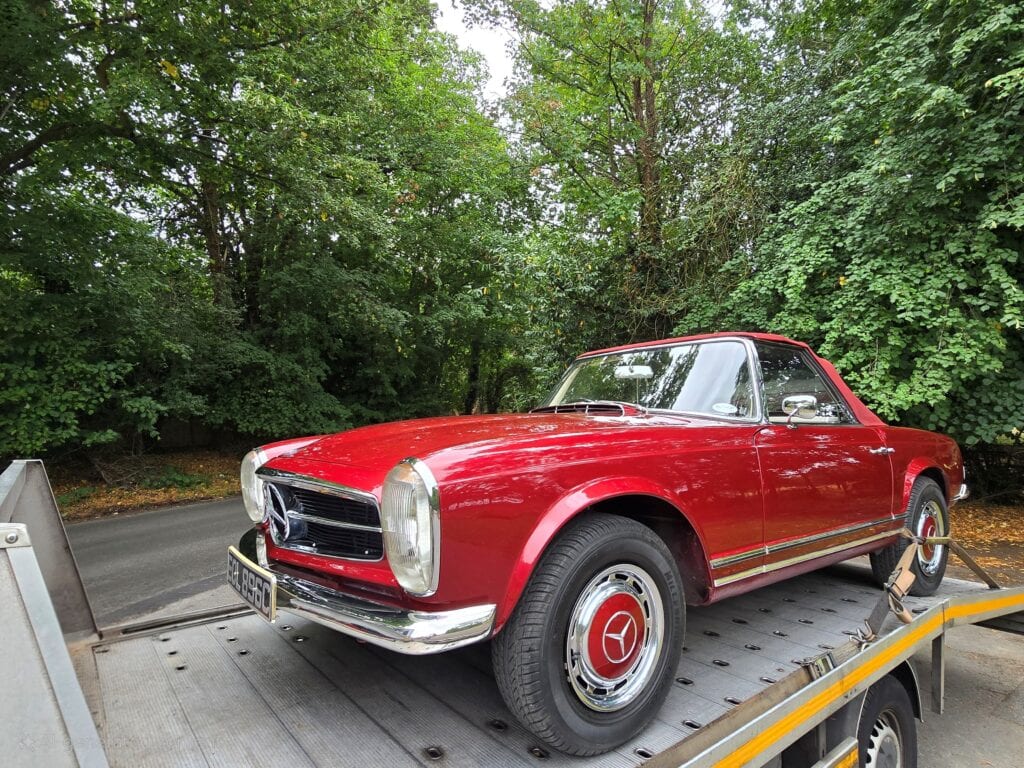 A classic red Mercedes-Benz SL convertible securely loaded onto a flatbed recovery truck on a tree-lined road in Surrey.