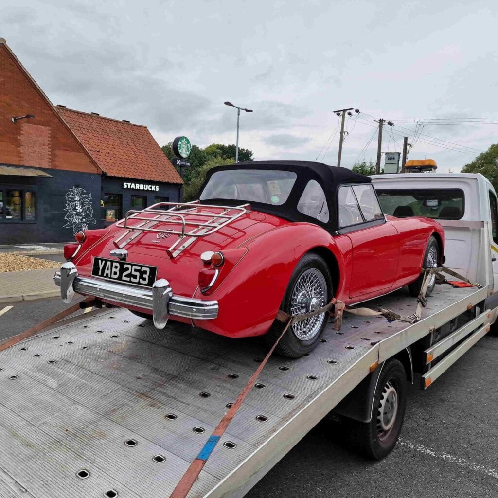 Rear view of a red classic MGA Roadster with a luggage rack securely loaded onto a flatbed truck, demonstrating safe vehicle transport.