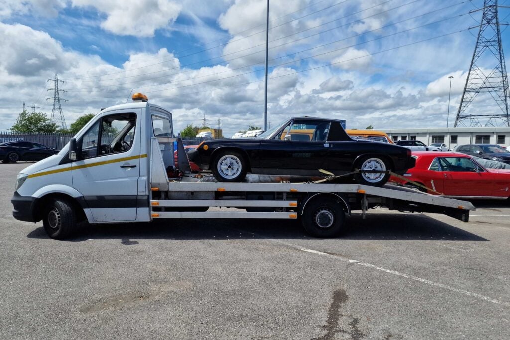 Side view of a black vintage Porsche 914 targa securely loaded onto a flatbed recovery truck in an industrial yard, showing safe vehicle transport.