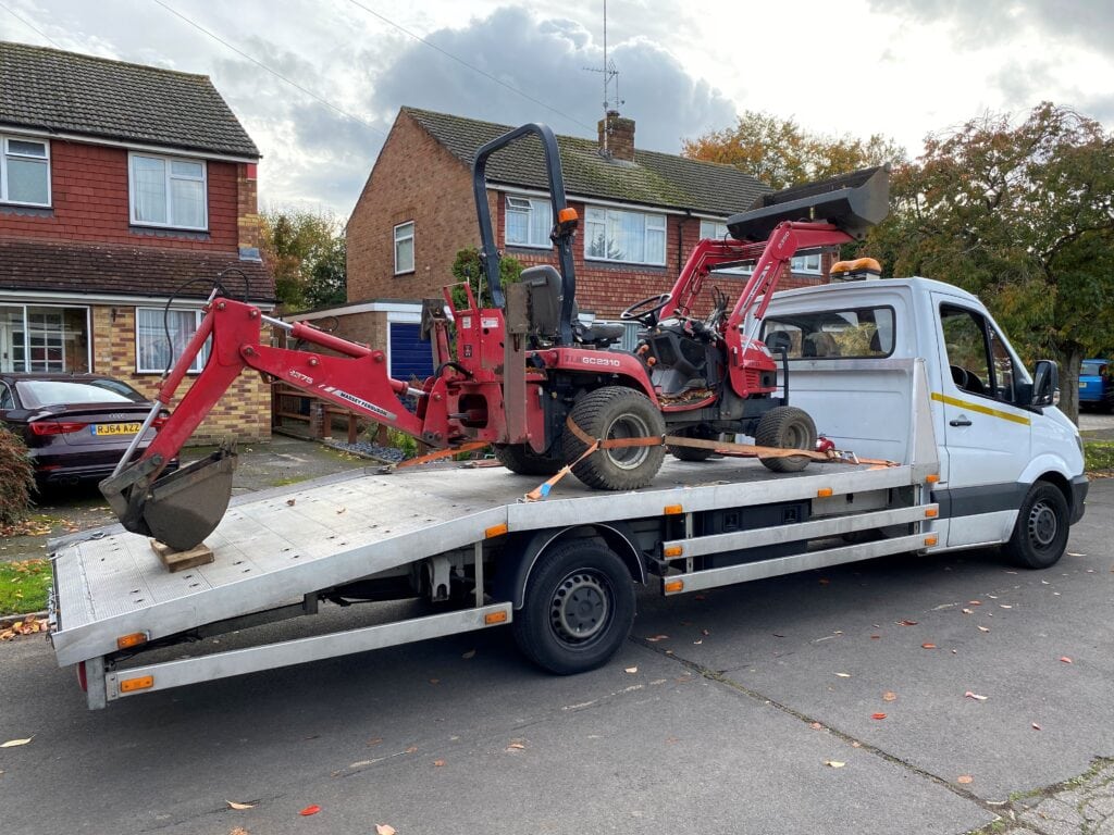 A small red Massey Ferguson compact tractor with a front loader and backhoe secured on a flatbed recovery truck in a residential street.