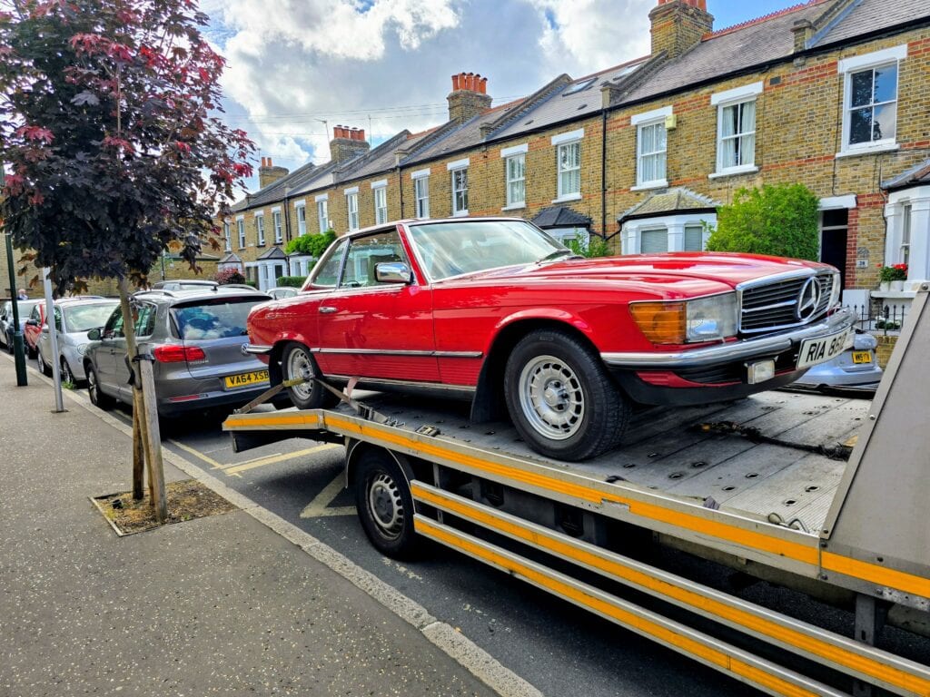 A classic red Mercedes-Benz SL roadster being loaded onto a flatbed recovery truck on a residential street in London.