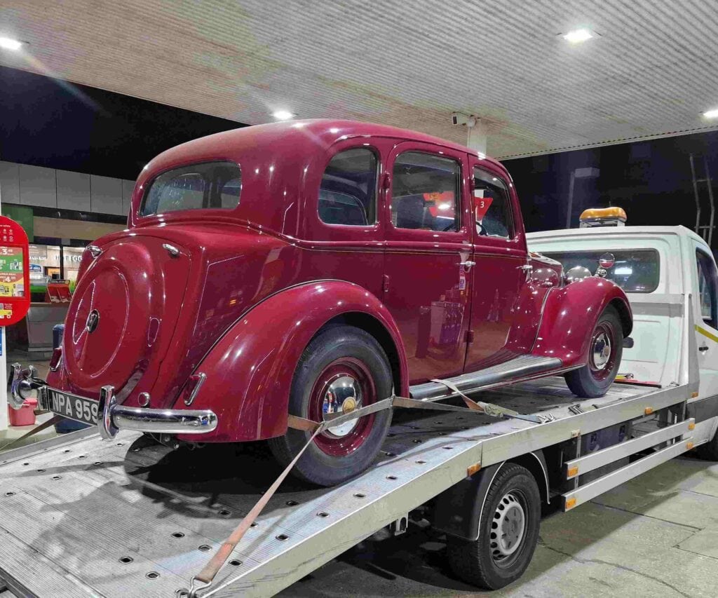 Rear view of a maroon vintage Riley RMA securely loaded on a flatbed recovery truck at a petrol station, demonstrating safe classic car transport.