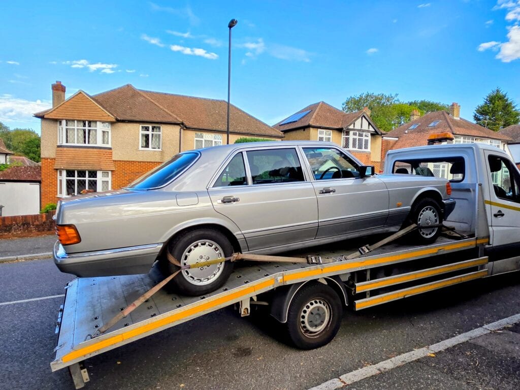 A silver Mercedes-Benz W126 S-Class being delivered on a flatbed recovery truck to a residential street in Surrey.
