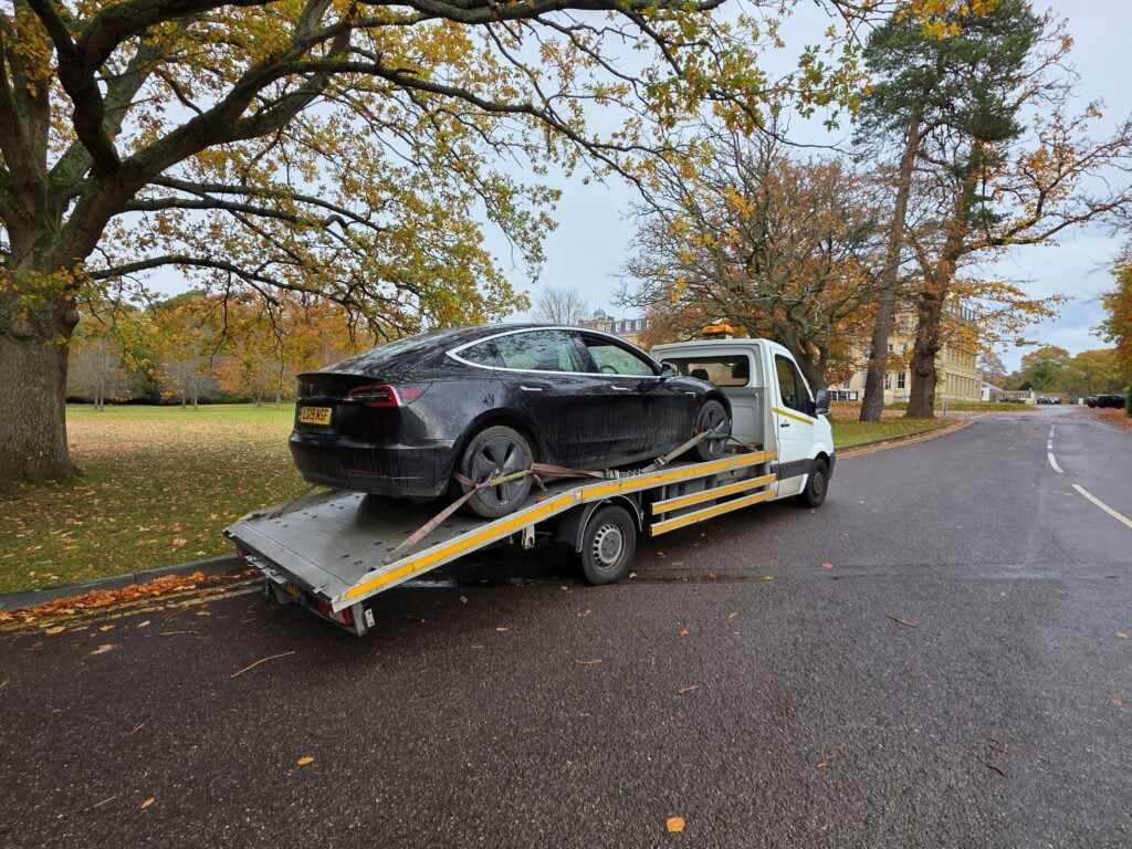 Black Tesla Model 3 loaded onto a recovery truck for emergency flatbed towing service in a park setting.