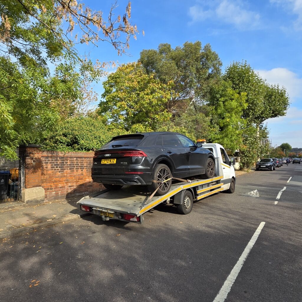 London Auto Towing flatbed recovery truck transporting a black Audi SUV on a residential street to demonstrate local car recovery service coverage.